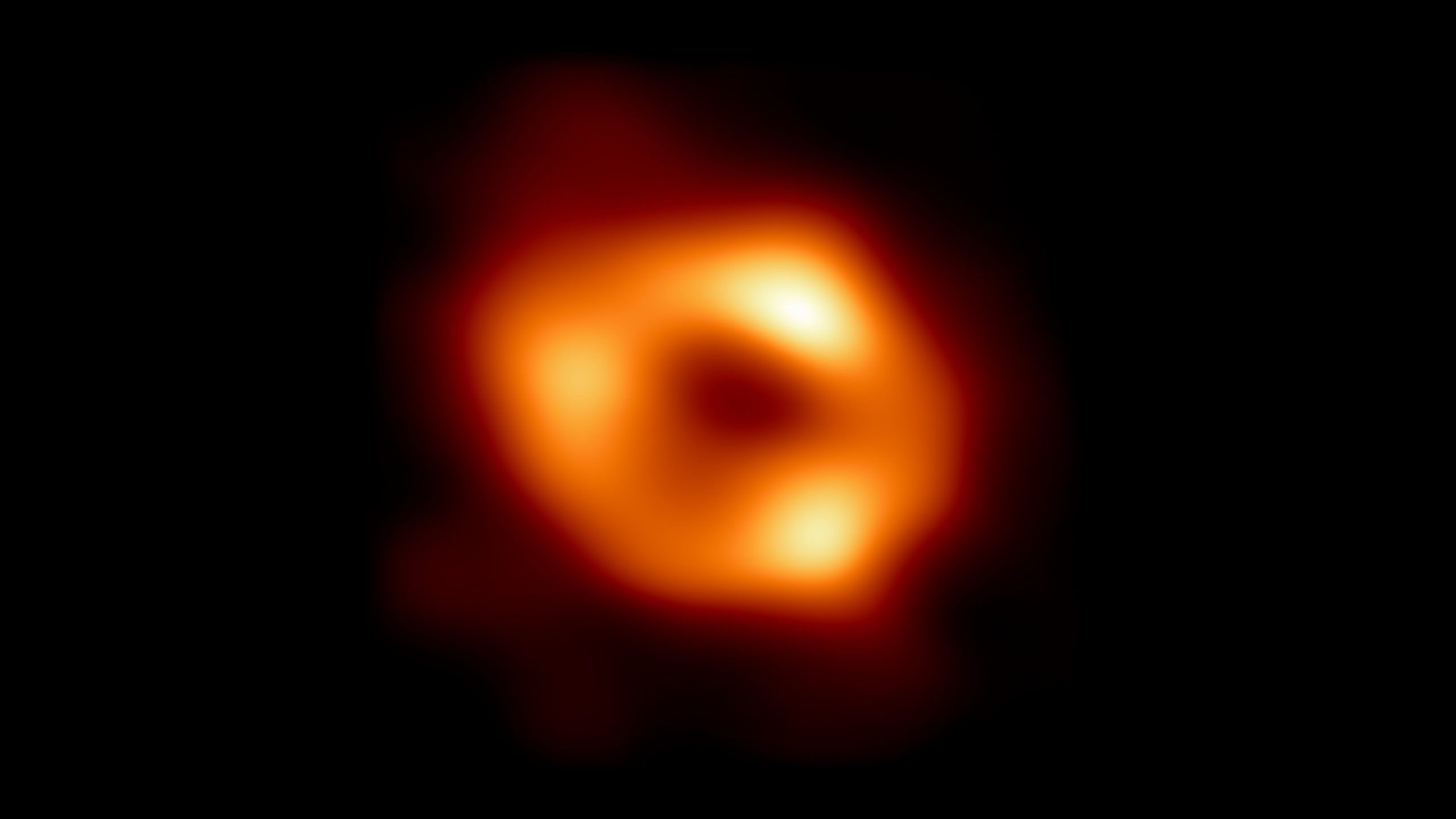 On top of the black background, the image shows a ring-like structure. This ring is made up of a diffuse transition of red, orange and yellow hues. The outer limbs of the ring are redder, depicting weaker emission. Inside of the ring, the colour is dark red, depicting the black hole shadow. On the ring, three brightness peaks reach yellow hues, depicting strong emission. The picture shows the first image of Sagittarius A*, the supermassive black hole at the centre of our galaxy. It is the first direct visual evidence of the presence of this black hole. It was captured by the Event Horizon Telescope (EHT), an array which linked together eight existing radio observatories across the planet to form a single “Earth-sized” virtual telescope. The telescope is named after the event horizon, the boundary of the black hole from inside of which no light can escape. Although we cannot see the event horizon itself, because it cannot emit light, glowing gas orbiting around the black hole reveals a telltale signature: a dark central region (called a shadow) surrounded by a bright ring-like structure. The new view captures light bent by the powerful gravity of the black hole, which is four million times more massive than our Sun. The image of the Sagittarius A* black hole is an average of the different images the EHT Collaboration has extracted from its 2017 observations.