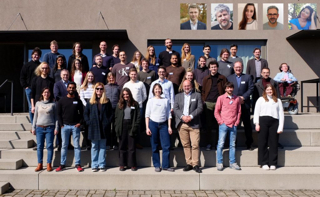 About 40 people are standing on a stairs in front of a building. Most people aare of age of approximately 30 or 40 years with few persons that are of age about 50 or 60. Three inset images show remote participants of the meeting.