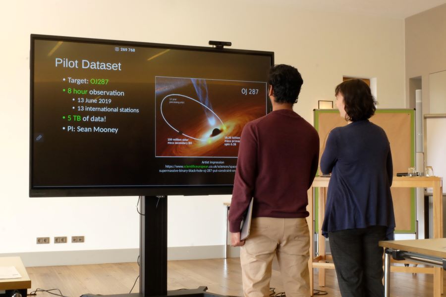 Decorative picture taken at the annual assembly 2023, showing two persons discussing in front of a screen displaying a presentation slide about the active galaxy OJ 287.